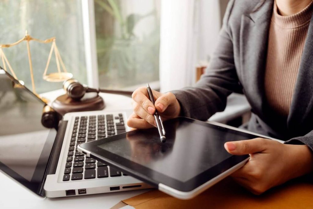 Profissional usando tablet em um escritório com laptop e elementos jurídicos ao fundo.