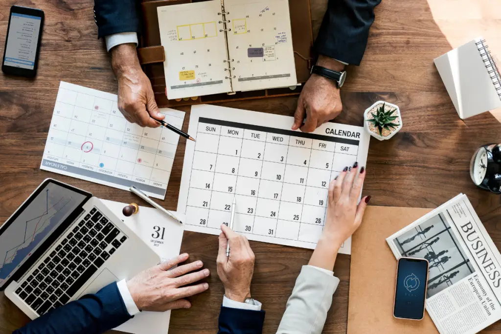 Mesa com um calendário no centro e mãos de alguns trabalhadores de terno sobre ele, com um notebook à esquerda representando software para controle de prazos judiciais.