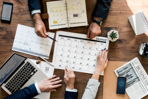 Mesa com um calendário no centro e mãos de alguns trabalhadores de terno sobre ele, com um notebook à esquerda representando software para controle de prazos judiciais.