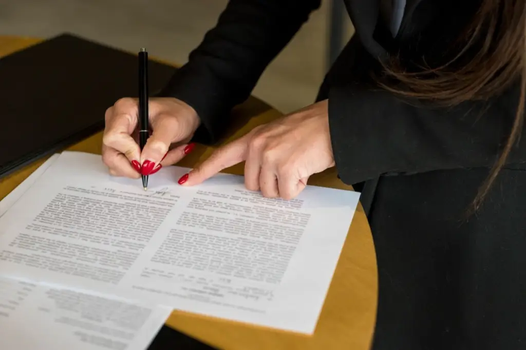 Mão feminina assinando contratos jurídicos sobre uma mesa de madeira.