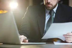 Homem de terno com um notebook aberto à esquerda, segurando documentos que representam um relatórios jurídicos.