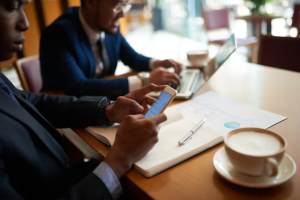 Homem negro organiza tarefas no celular, com um colaborador ao lado e uma xícara de café sobre a mesa, ilustrando um conteúdo do Gojur sobre gestão de tarefas na advocacia.