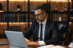 Um advogado em um escritório com estante repleta de livros, digita em um notebook atento, ilustrando um artigo do GOJUR sobre como ser um bom advogado.